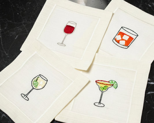 A variety of cream cotton napkins featuring embroidered icons of a Pina Colada, Gin & Tonic, and Aperol Spritz.
