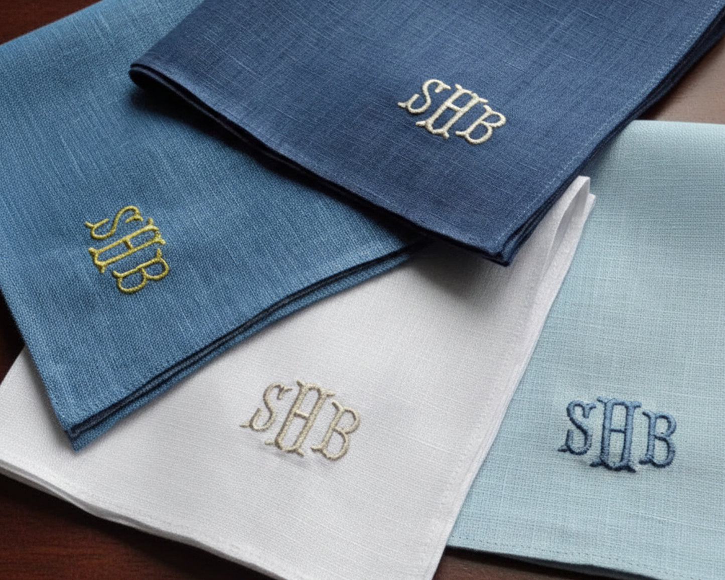 A collection of four linen pocket squares in navy, blue, and white with different thread color monograms.