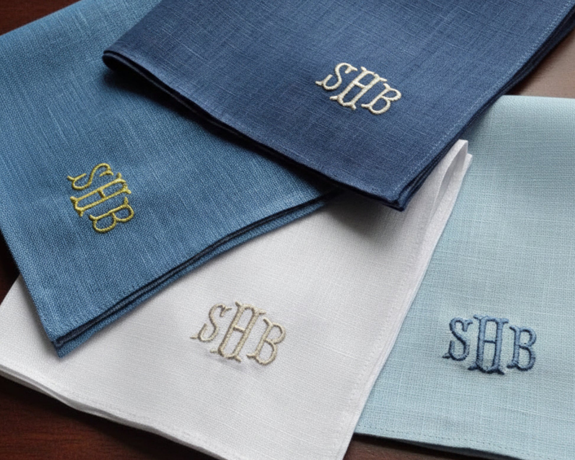 A collection of four linen pocket squares in navy, blue, and white with different thread color monograms.
