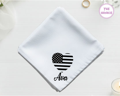 A crisp white cotton handkerchief personalized with the name "Ava" below a black-threaded heart containing American flag stars and stripes.