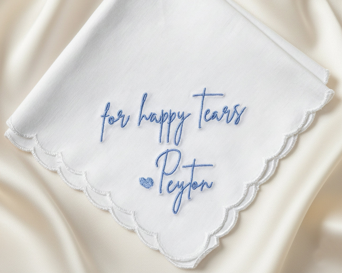 A close-up of a scalloped-edge wedding handkerchief showcasing intricate blue machine embroidery for personalized "Happy Tears" script.