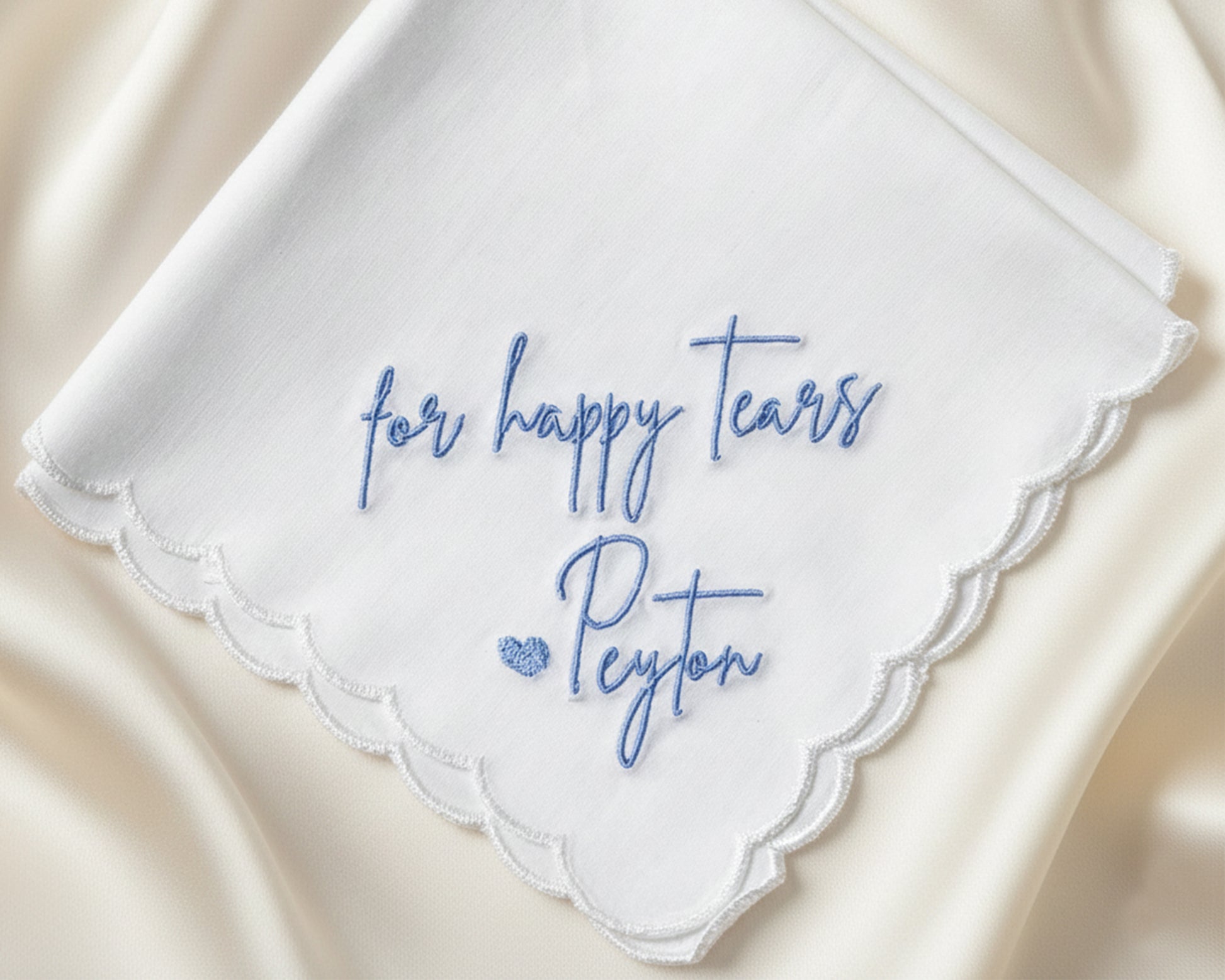 A close-up of a scalloped-edge wedding handkerchief showcasing intricate blue machine embroidery for personalized "Happy Tears" script.