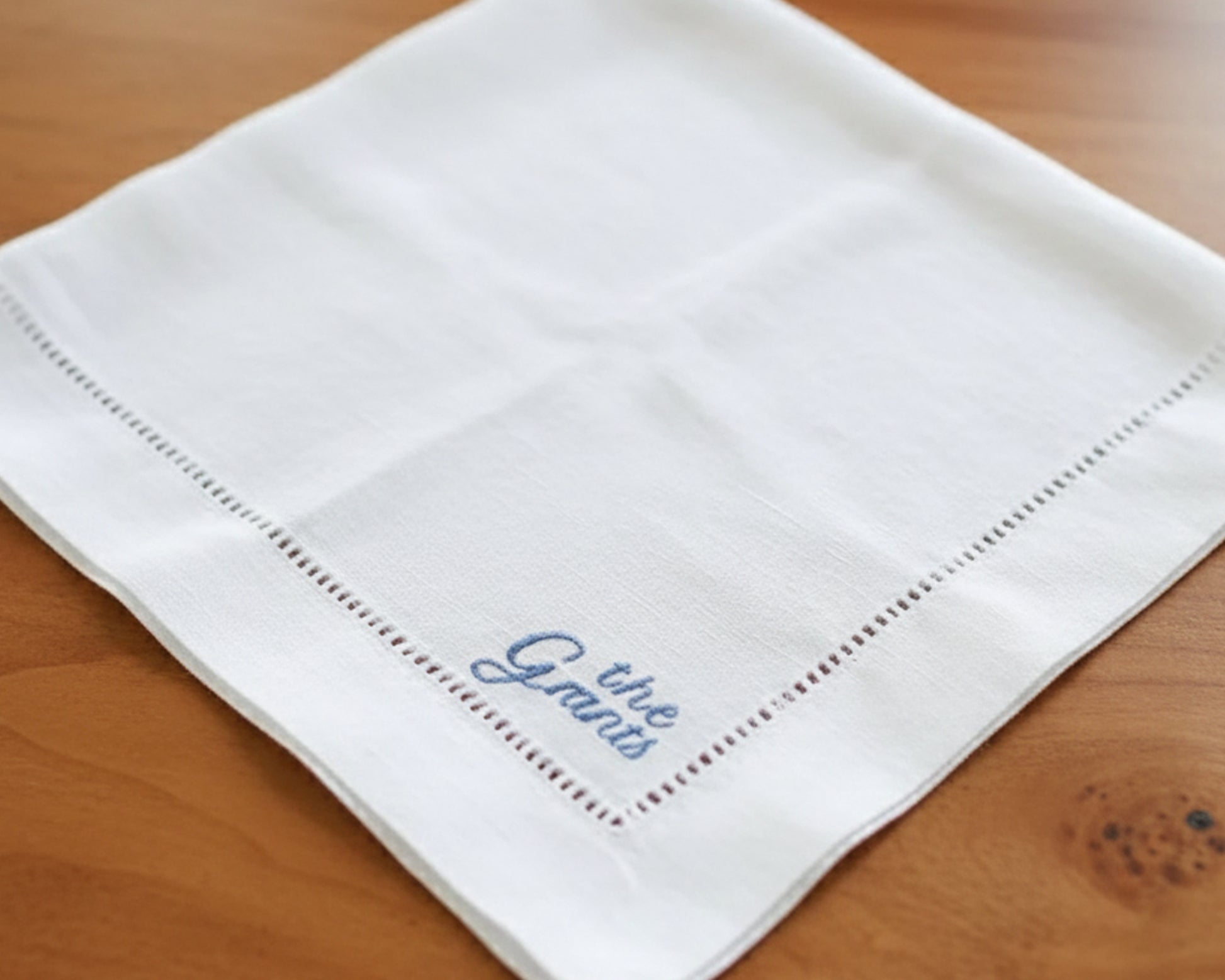 A collection of white personalized hemstitch napkins fanned out on a white wood surface, showcasing the elegant script and classic border.