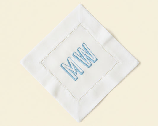 Single white cotton cocktail napkin featuring a large navy blue custom embroidered initial with a decorative hemstitch border.