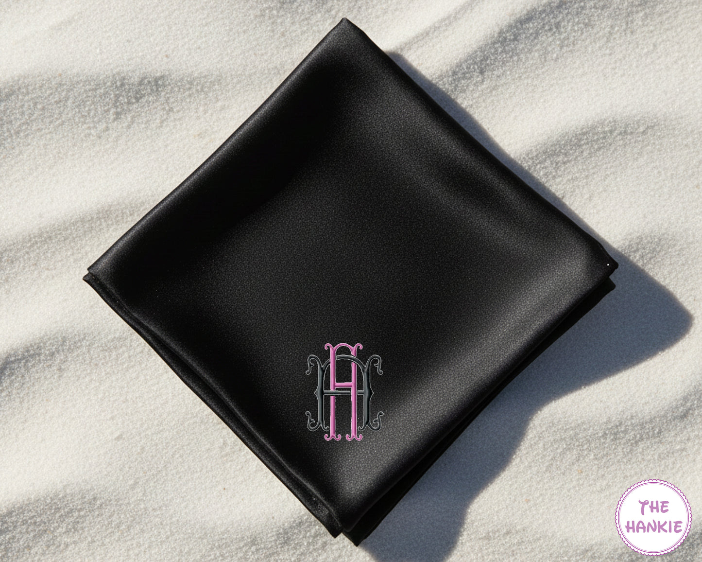 A bespoke black satin handkerchief with a classic royal blue embroidered monogram, shown alongside sunglasses and sunscreen on sand.