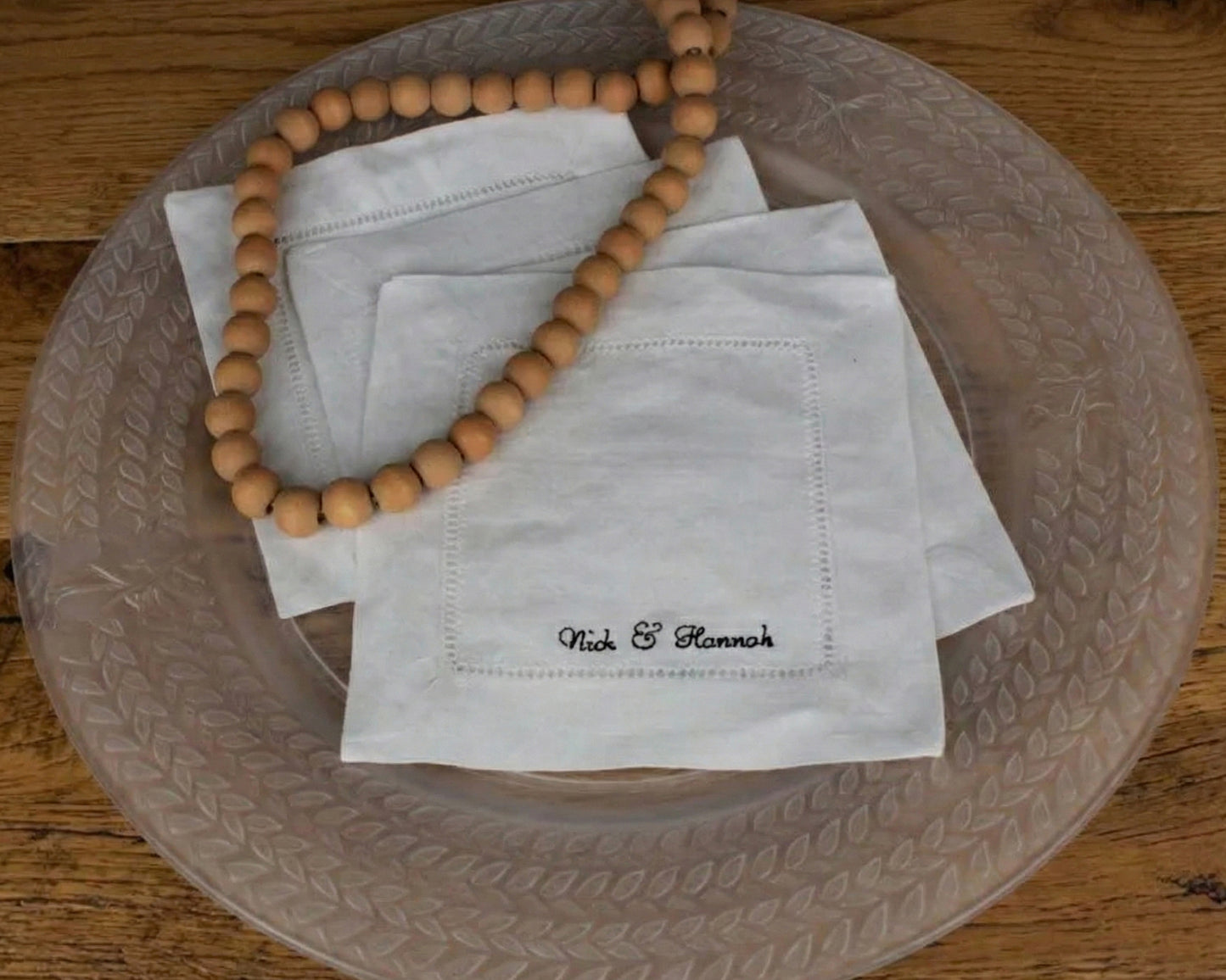 A stack of three personalized white linen hemstitch napkins featuring custom name embroidery, styled with a wooden bead garland on a glass plate.