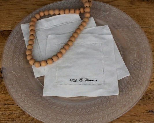 A stack of three personalized white linen hemstitch napkins featuring custom name embroidery, styled with a wooden bead garland on a glass plate.