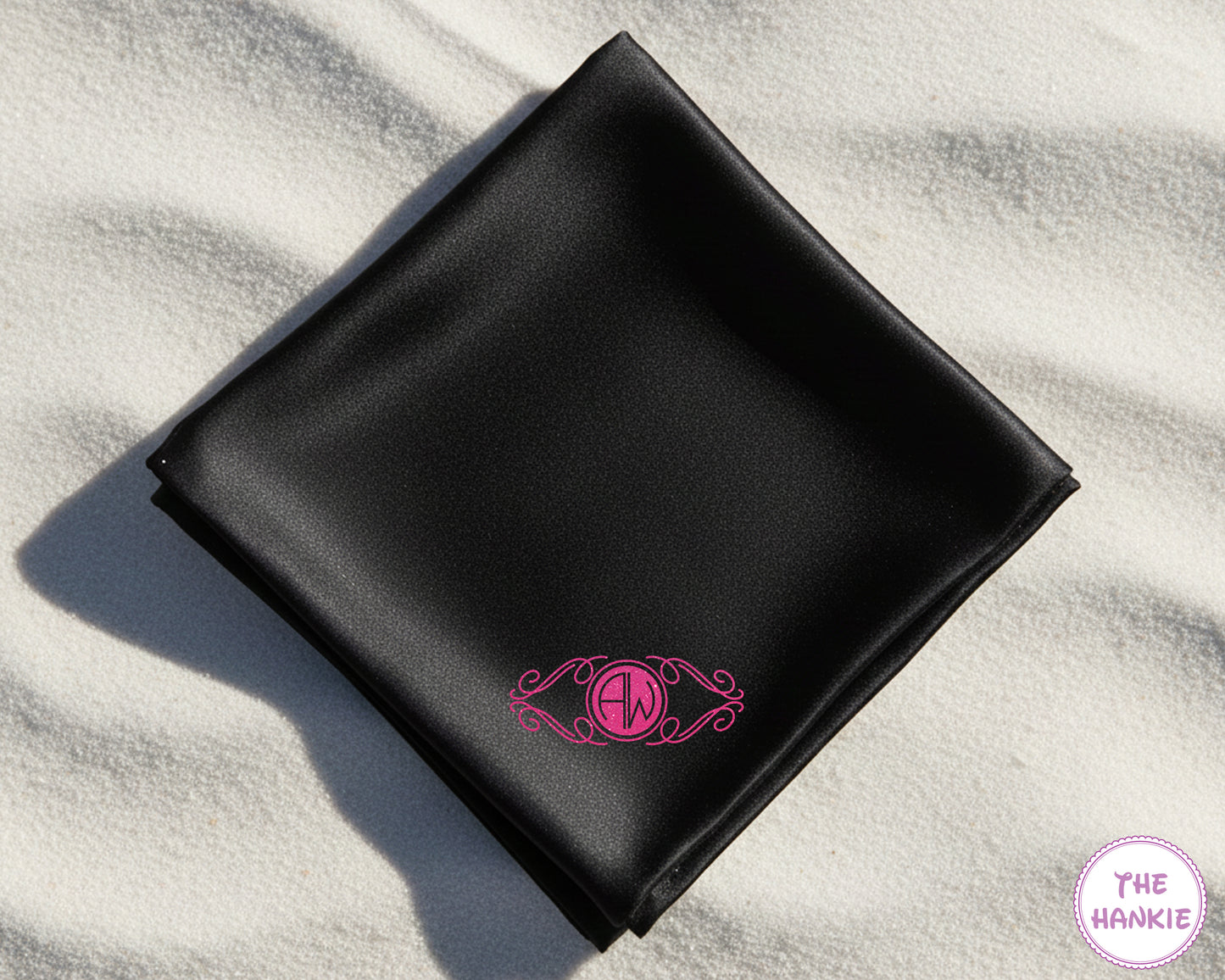 A black luxury satin handkerchief featuring a custom vibrant pink vinyl "AA" monogram on a white sand beach.