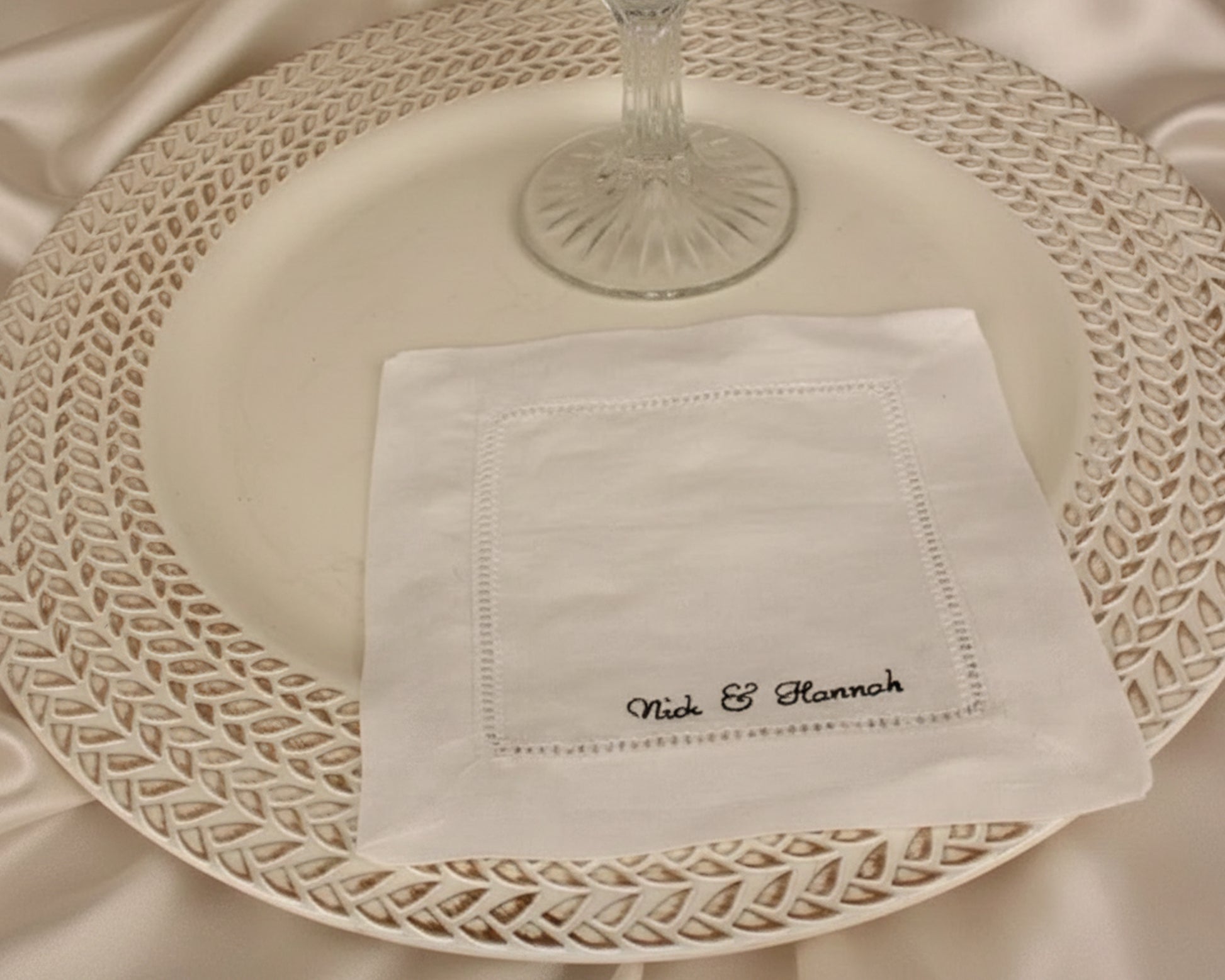 A crisp white linen cocktail napkin with a black-highlighted hemstitch border and custom "Nick & Hannah" embroidery on a dark marble surface.
