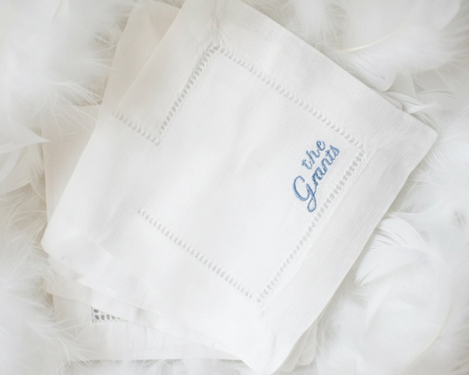 A stack of three premium white cotton napkins with hemstitch detailing and custom blue script embroidery displayed on a dark surface.