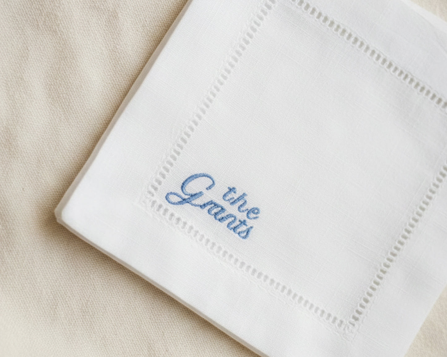 A luxury white gift box containing a personalized hemstitch cotton napkin with sky blue script embroidery, secured with a white ribbon.