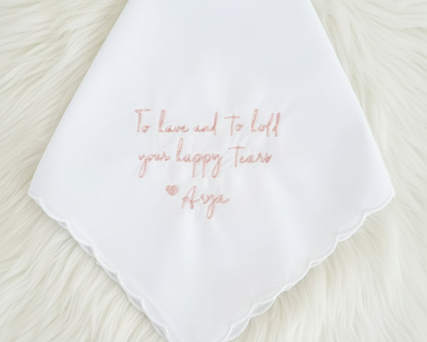 A white cotton handkerchief with a scalloped edge and "To have and to hold your happy tears Arya" in soft pink embroidery.