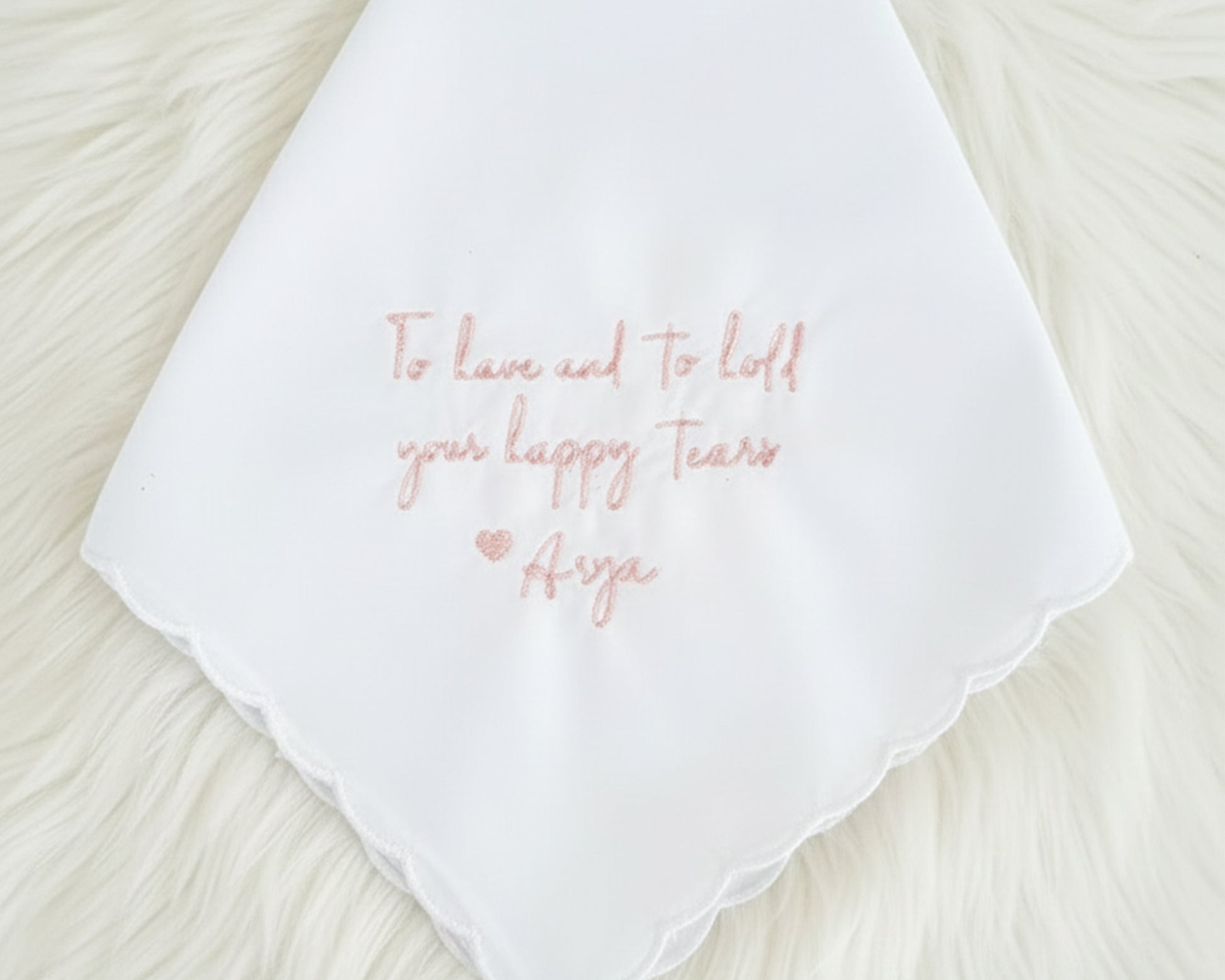 A white cotton handkerchief with a scalloped edge and "To have and to hold your happy tears Arya" in soft pink embroidery.
