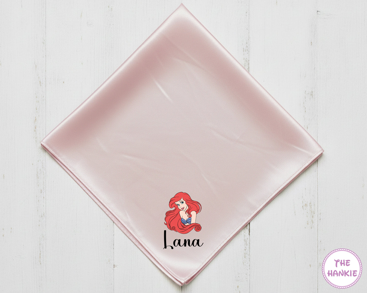 Pink satin square with a character design and text on a white wooden surface, branded 'The Hankie'.