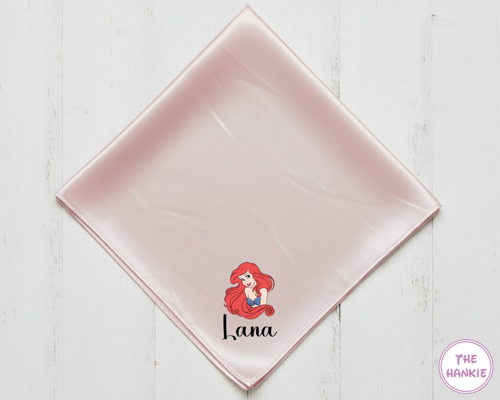 Pink satin square with a character design and text on a white wooden surface, branded 'The Hankie'.