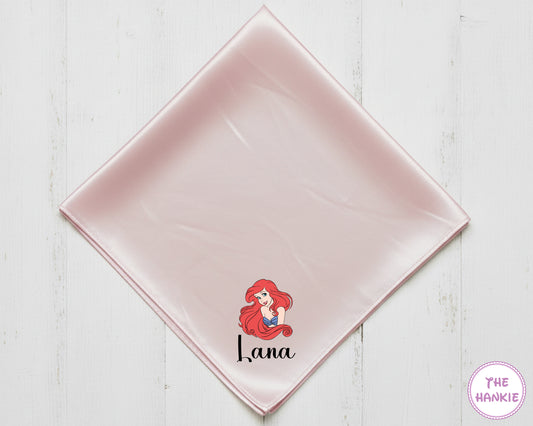 Pink satin square with a character design and text on a white wooden surface, branded 'The Hankie'.