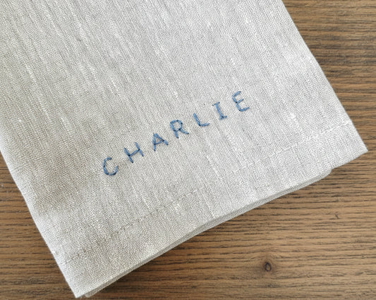 A close-up of a natural-colored cotton linen napkin featuring the name "CHARLIE" hand-embroidered in a slate blue textured thread.