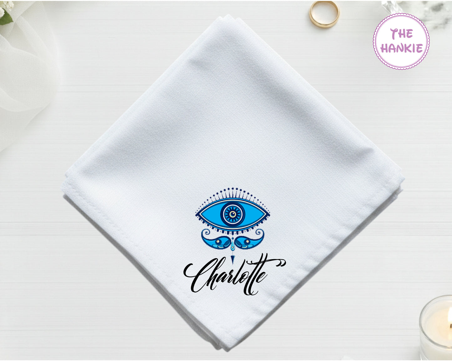 A crisp white cotton handkerchief personalized with "Charlotte" below an intricate blue and black bohemian-style evil eye with detailed eyelashes.