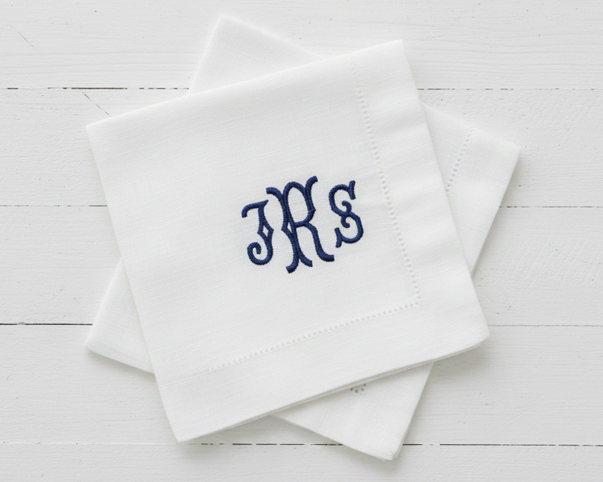 A stack of two white linen cocktail napkins featuring a classic navy blue embroidered monogram in a serif font.