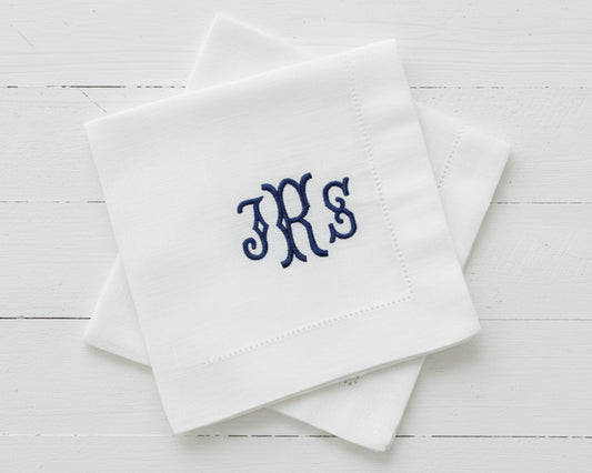 A stack of two white linen cocktail napkins featuring a classic navy blue embroidered monogram in a serif font.