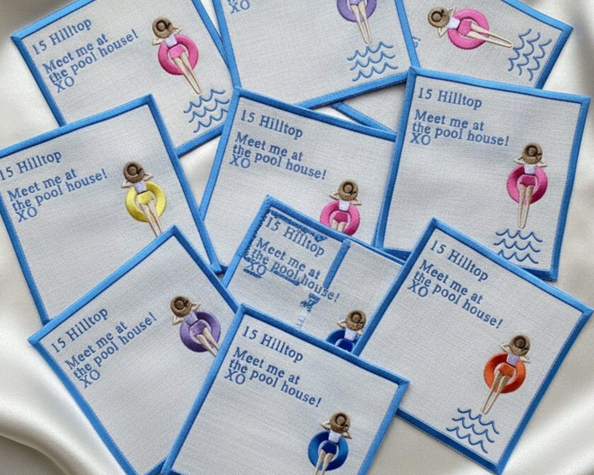 A fan-style display of personalized pool house napkins with blue borders and custom address embroidery on a satin background.