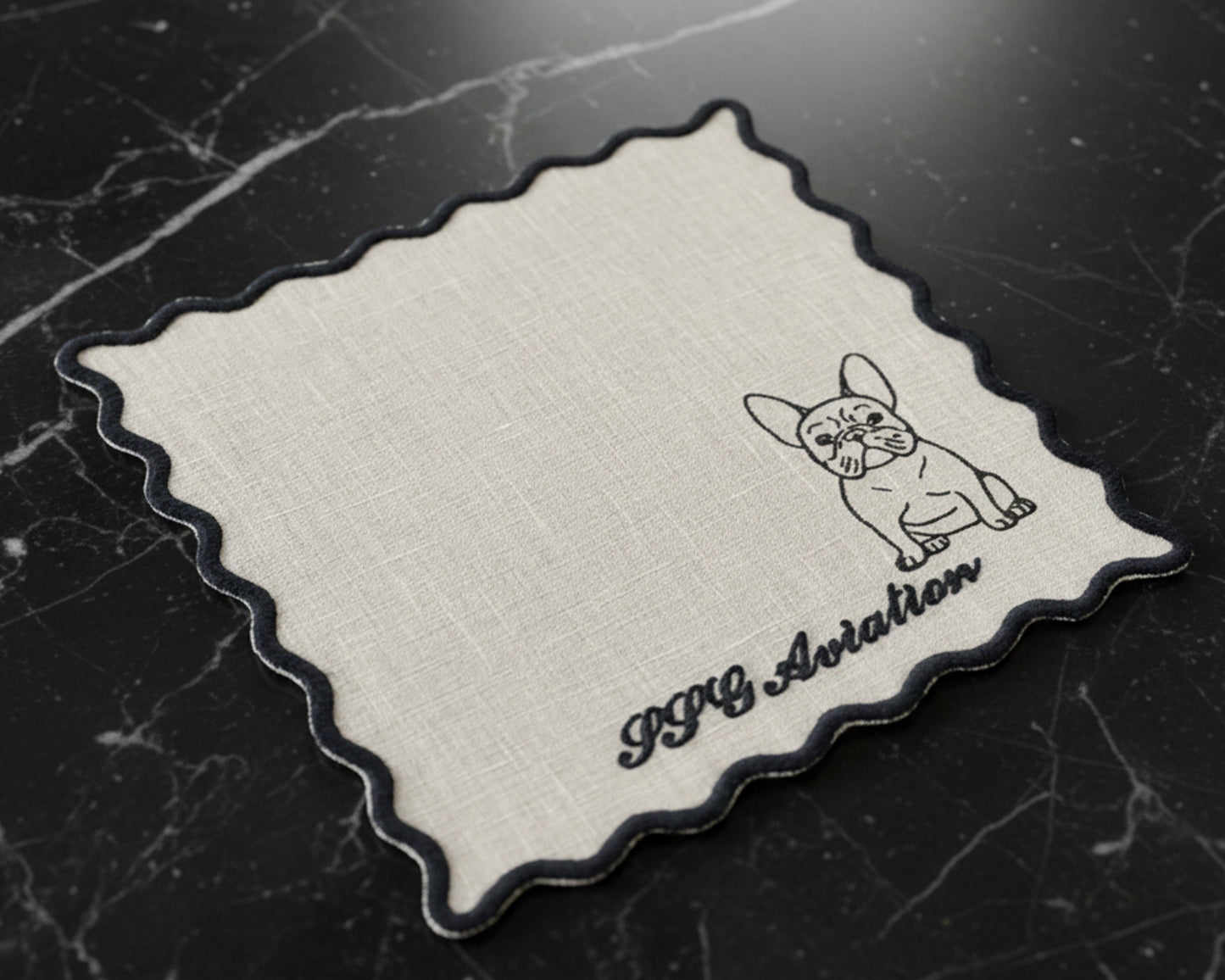 A beige linen cocktail napkin with a black scalloped edge and "PPG Aviation" logo embroidery on a black marble surface.