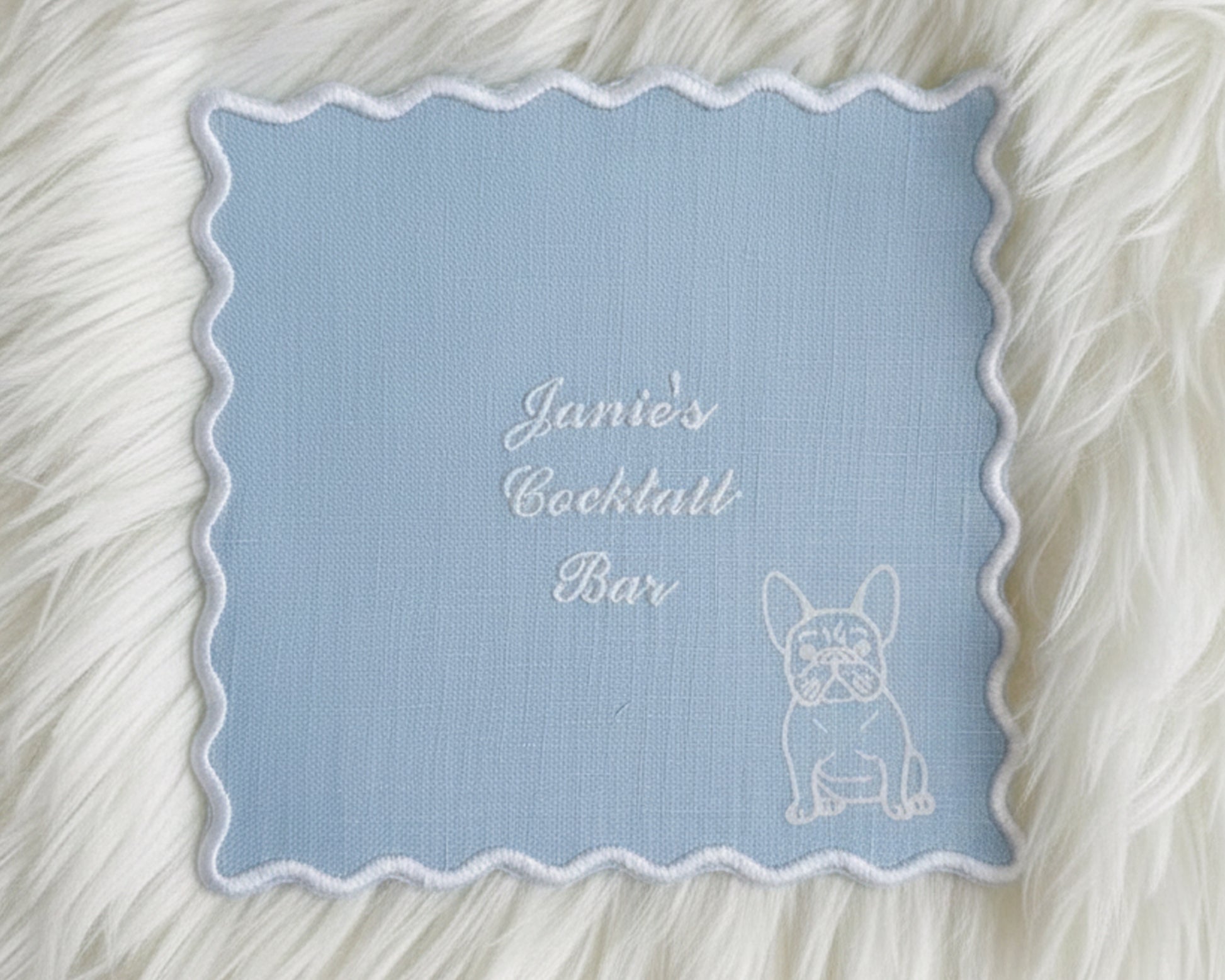 A light blue linen cocktail napkin with a white scalloped edge, embroidered with "Janie’s Cocktail Bar" and a white French Bulldog illustration.