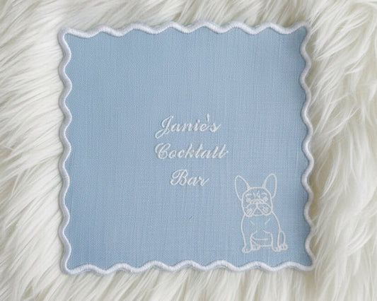 A light blue linen cocktail napkin with a white scalloped edge, embroidered with "Janie’s Cocktail Bar" and a white French Bulldog illustration.