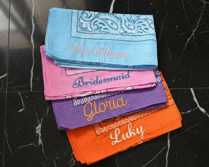 Four personalized bachelorette bandanas with script embroidery displayed on a soft white feather background.