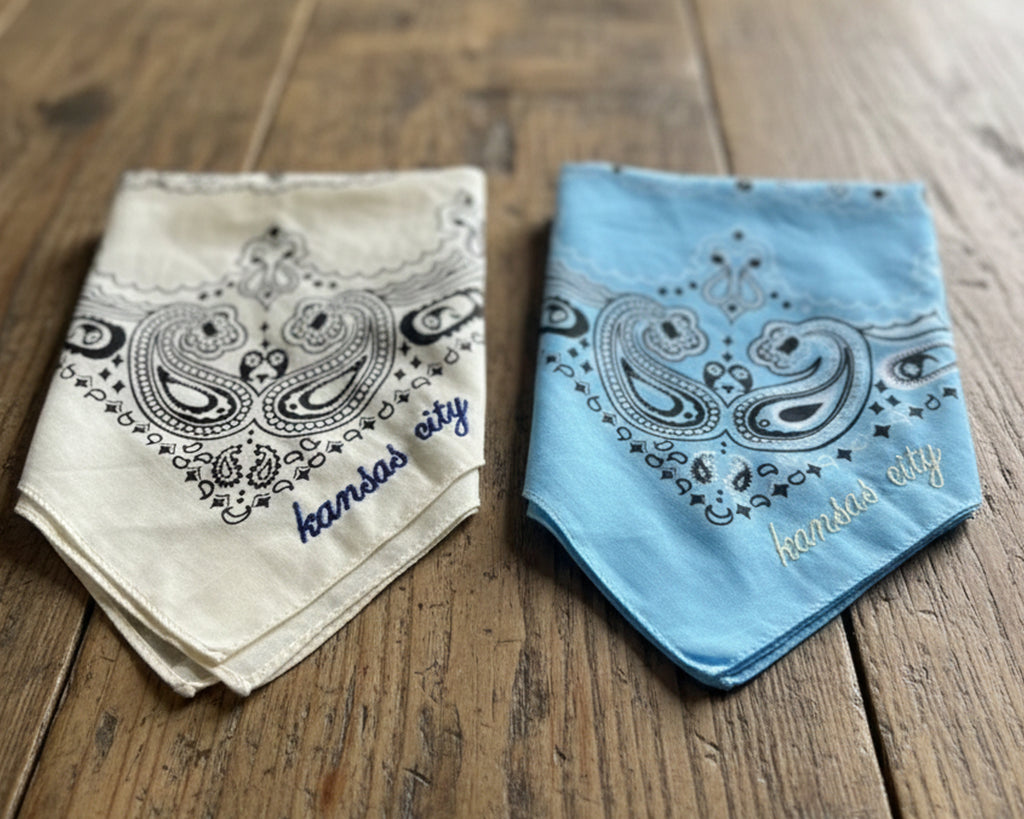 A light-colored paisley bandana with custom red chain stitch embroidery, styled on a wooden surface with a western hat and turquoise jewelry.