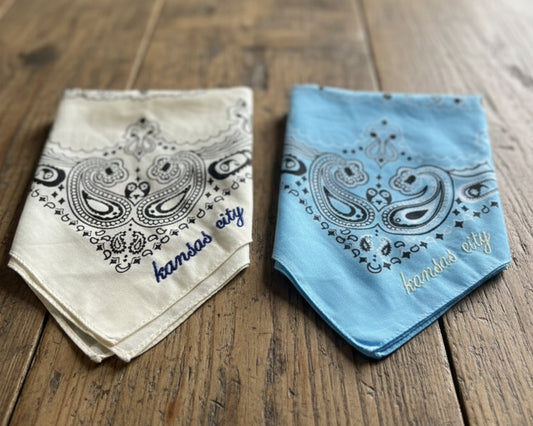 A light-colored paisley bandana with custom red chain stitch embroidery, styled on a wooden surface with a western hat and turquoise jewelry.