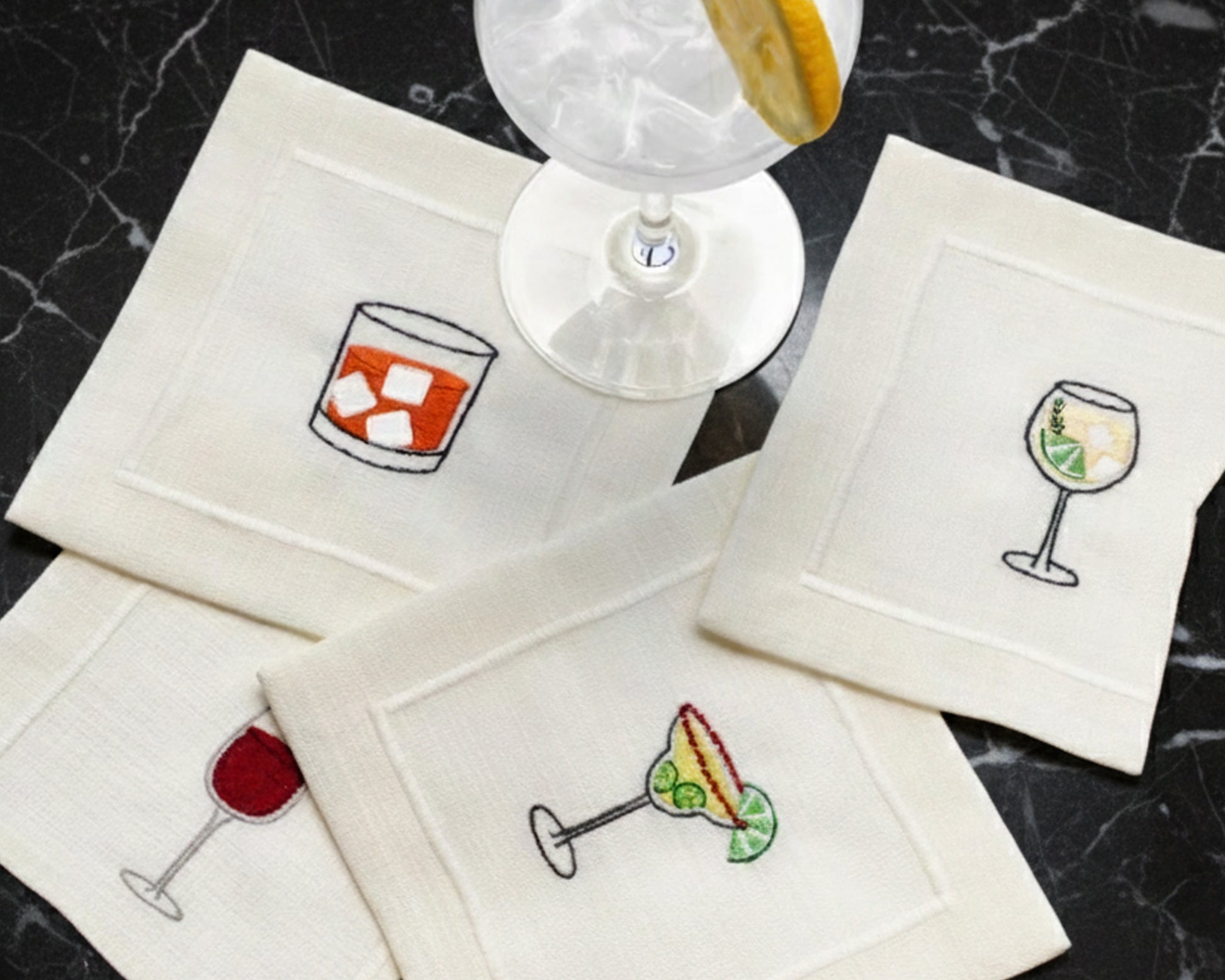 Four cream cotton cocktail napkins with embroidered drink icons, including an Old Fashioned, red wine, and Margarita, displayed on a black marble surface next to a cocktail glass.