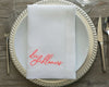 A personalized white linen napkin with coral embroidery placed on a silver-rimmed charger plate with formal cutlery