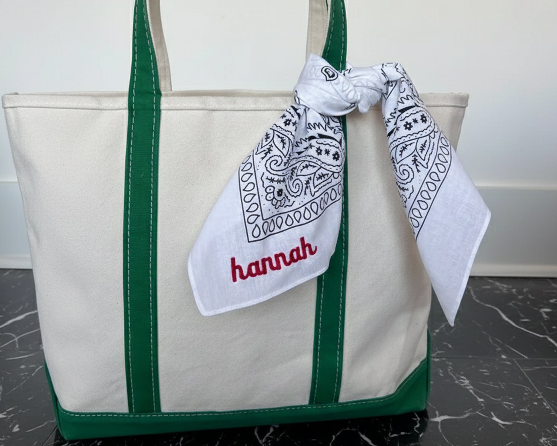 A white paisley bandana with the name "hannah" embroidered in red script, tied to the handle of a cream and green canvas tote bag.