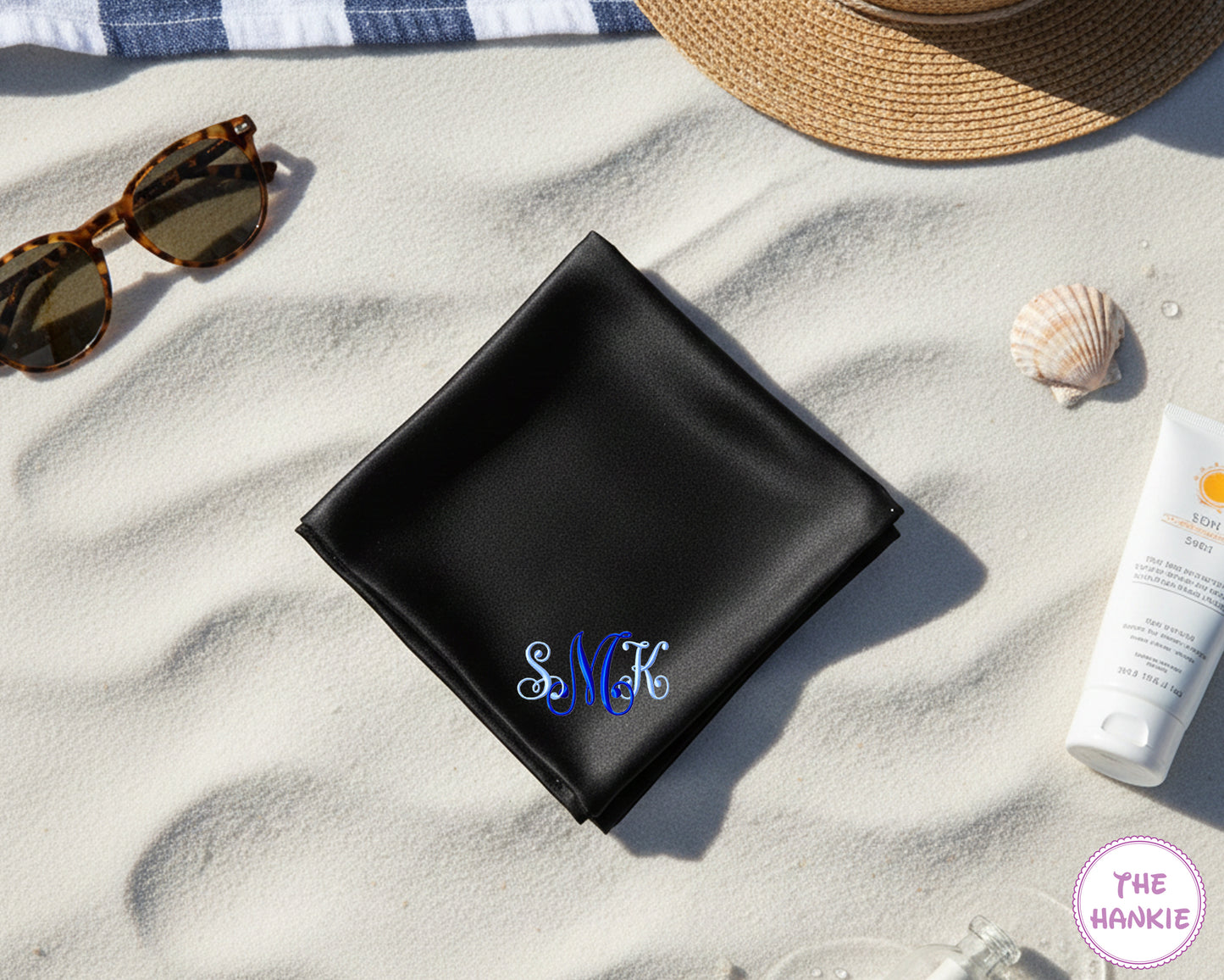 A bespoke black satin handkerchief showcasing a classic blue embroidered monogram with decorative swirls, displayed in a beach setting.
