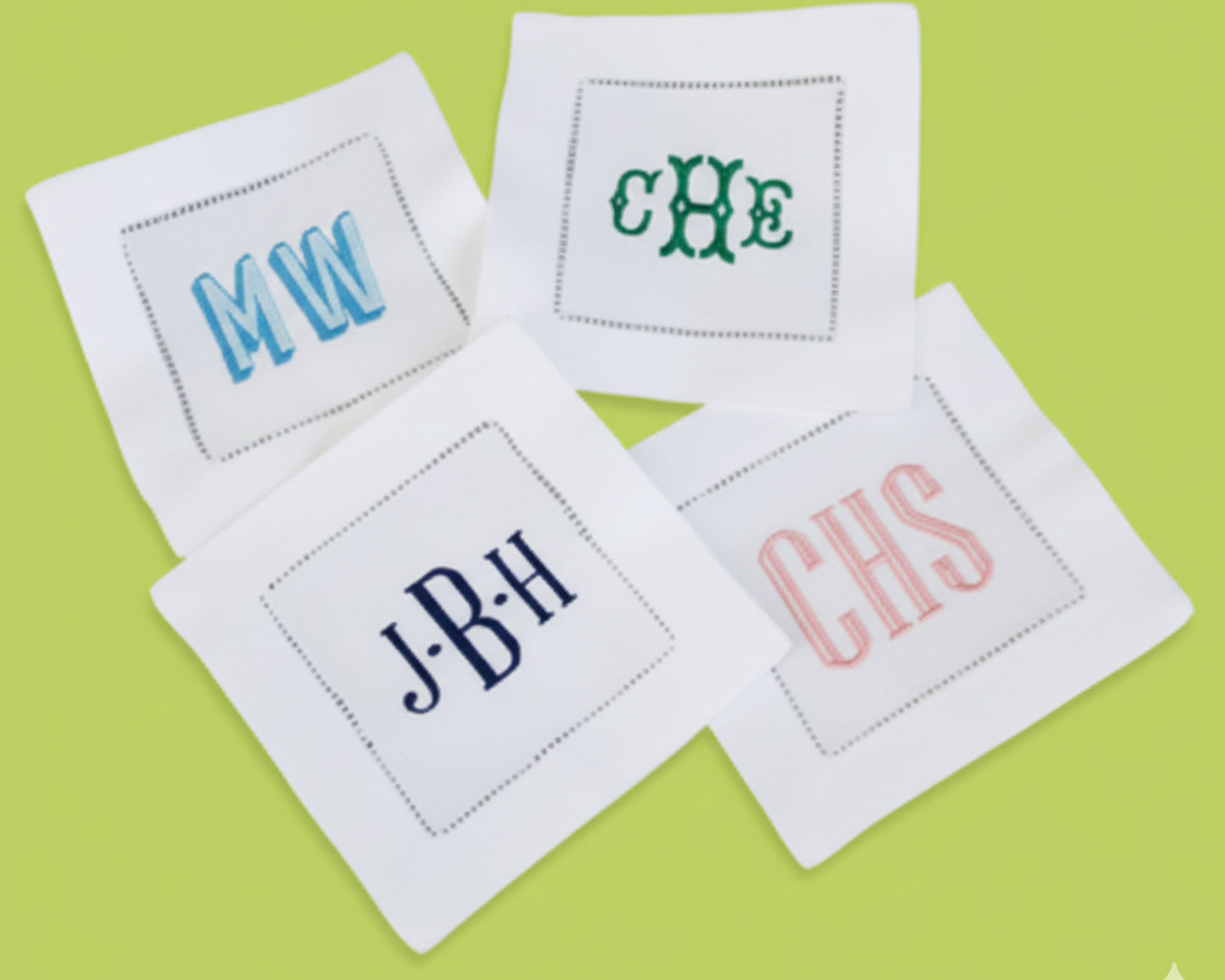 A set of four white linen napkins featuring different custom monogram embroidery styles in blue, green, and pink.