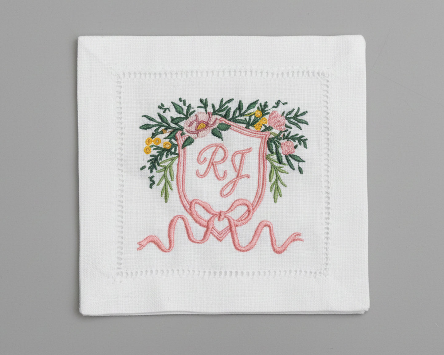 White hemstitch napkin featuring a detailed pink floral heraldic crest with custom 'RJ' initials