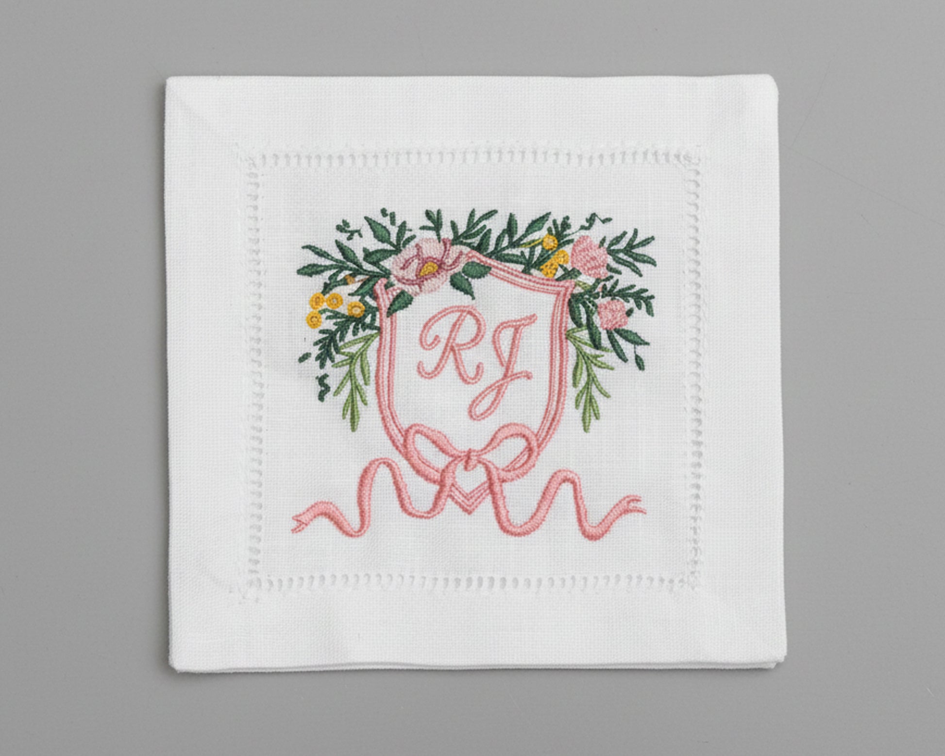White hemstitch napkin featuring a detailed pink floral heraldic crest with custom 'RJ' initials