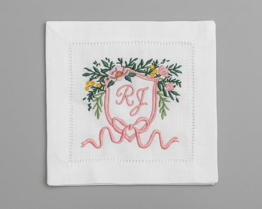 White hemstitch napkin featuring a detailed pink floral heraldic crest with custom 'RJ' initials