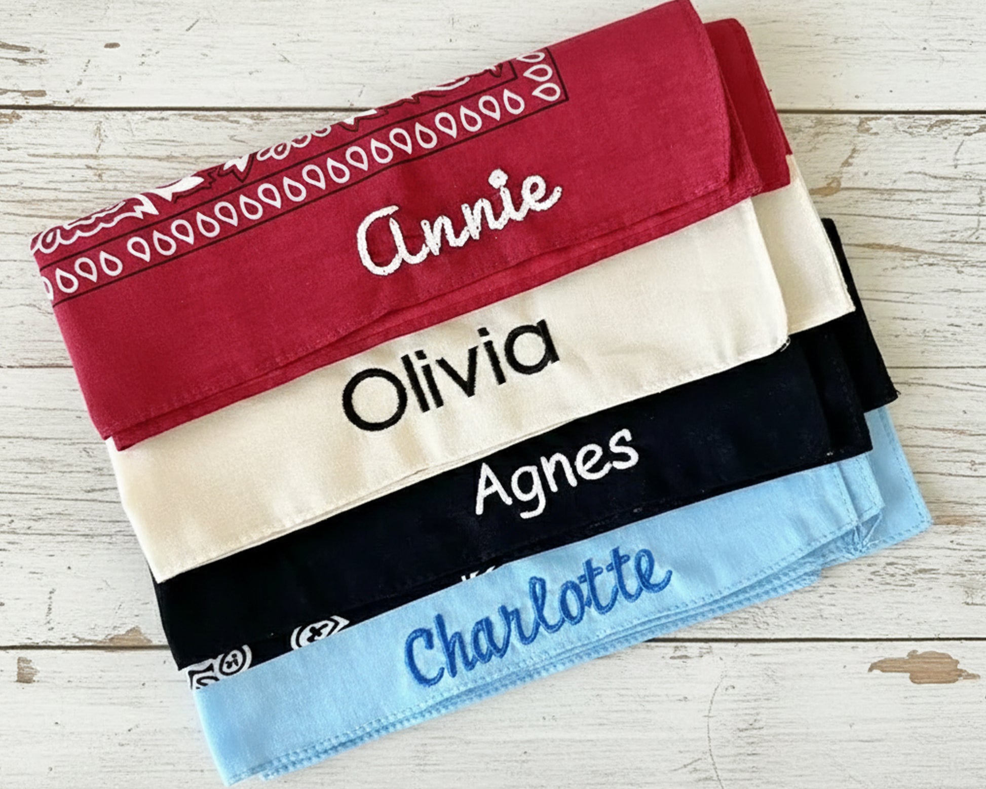 A close-up view of a folded burgundy red paisley bandana with the name "Annie" embroidered in white chain stitch script