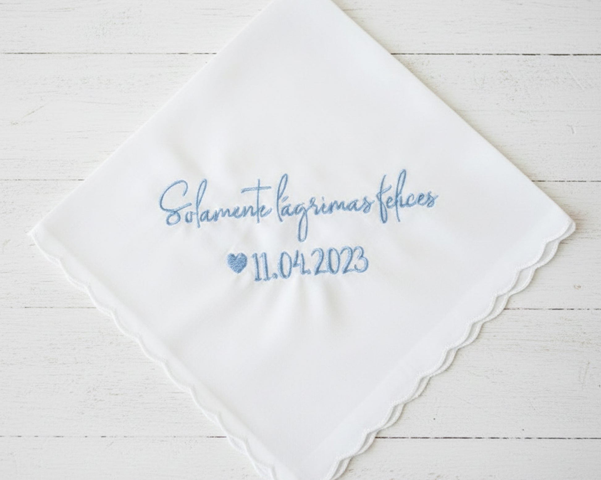 Personalized Spanish wedding handkerchief with a heart and the phrase "Solamente lágrimas felices" in light blue embroidery, shown on soft pink silk.