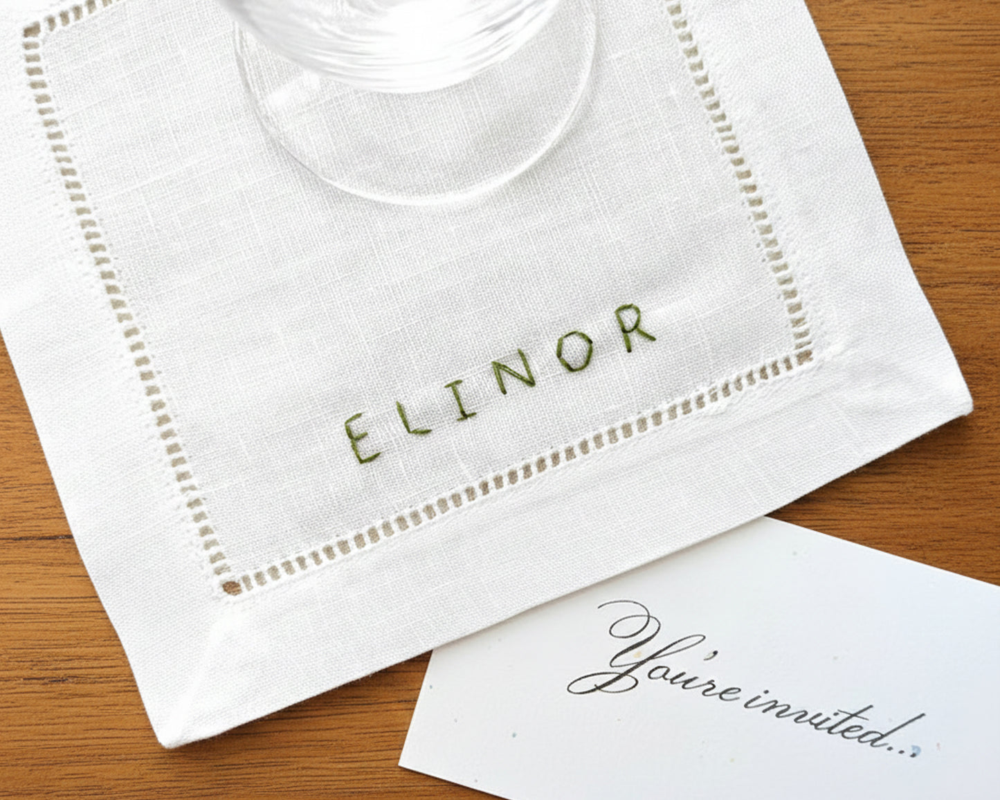 A white cotton hemstitch napkin featuring "ELINOR" hand-stitched in a moss green thread, placed next to a "You're invited" card.