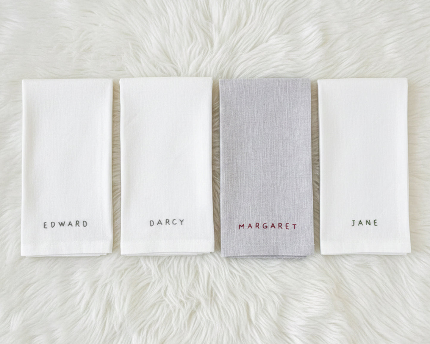 A white cotton beverage napkin with a perfectly straight machine-hemstitch border, featuring the name "ELINOR" in moss green digital embroidery.