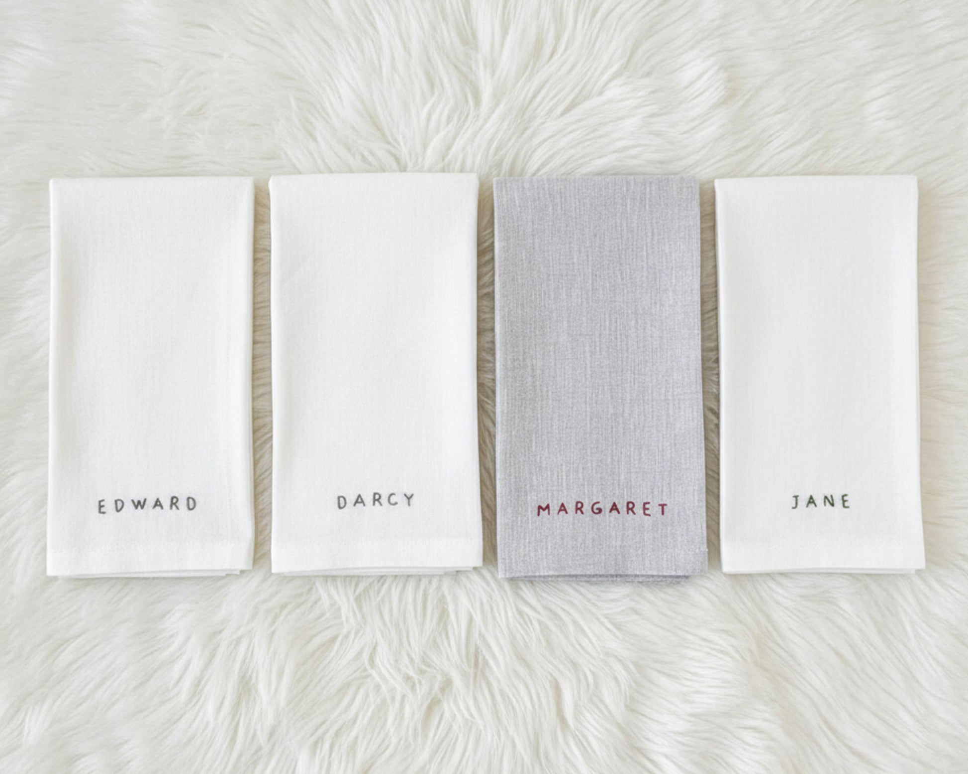 A white cotton beverage napkin with a perfectly straight machine-hemstitch border, featuring the name "ELINOR" in moss green digital embroidery.