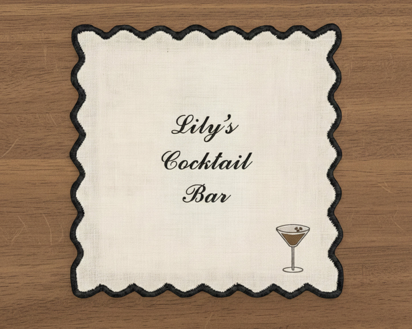 A white cotton cocktail napkin with a sharp black wave-edge border, featuring personalized script and a dark espresso martini icon.