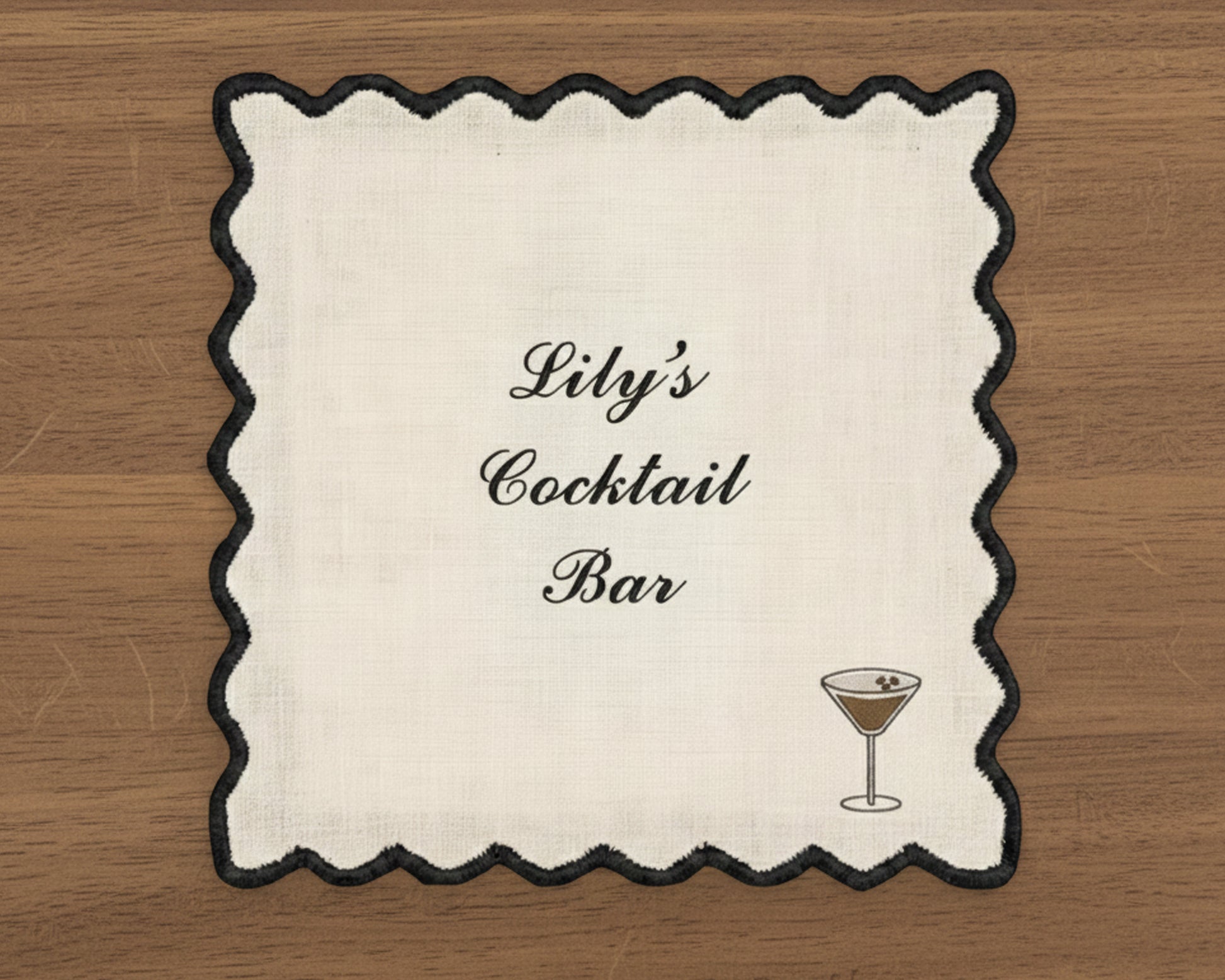 A white cotton cocktail napkin with a sharp black wave-edge border, featuring personalized script and a dark espresso martini icon.