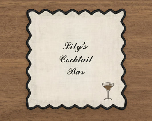 A white cotton cocktail napkin with a sharp black wave-edge border, featuring personalized script and a dark espresso martini icon.