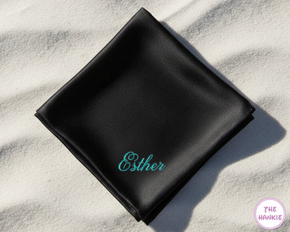 A premium black satin pocket square with the name "Esther" personalized in a shimmering teal script embroidery.