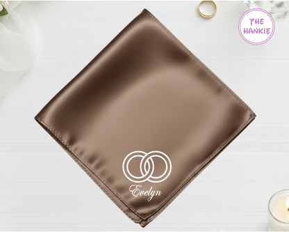A burnished bronze high-lustre satin pocket square customized with "Evelyn" and white embroidery of two interlocking wedding bands.