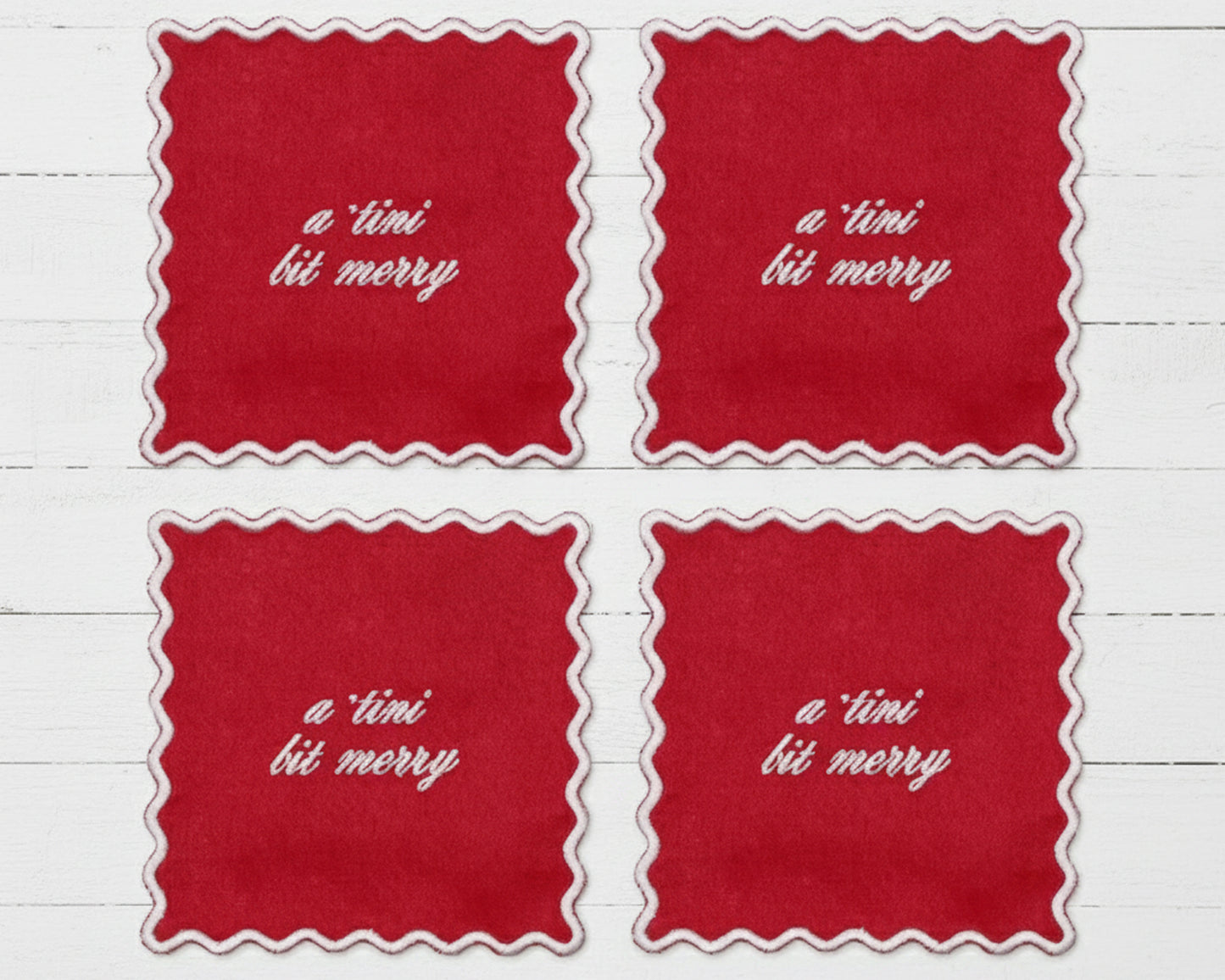 A set of four ruby red linen cocktail napkins with white scalloped edges featuring "a tini bit merry" in white vinyl script.